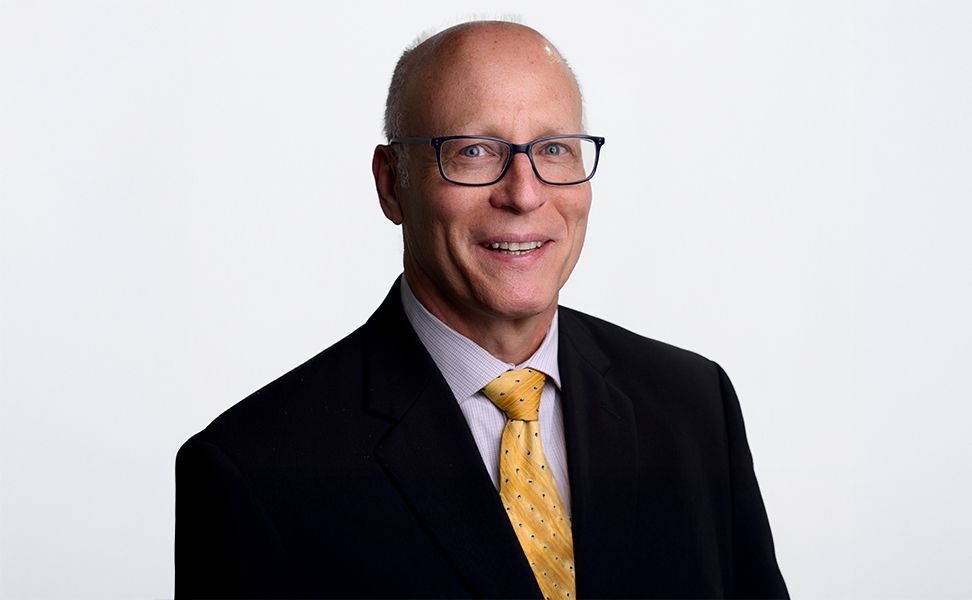 Joseph Turner wearing glasses, a suit, and a yellow tie smiles against a plain white background.