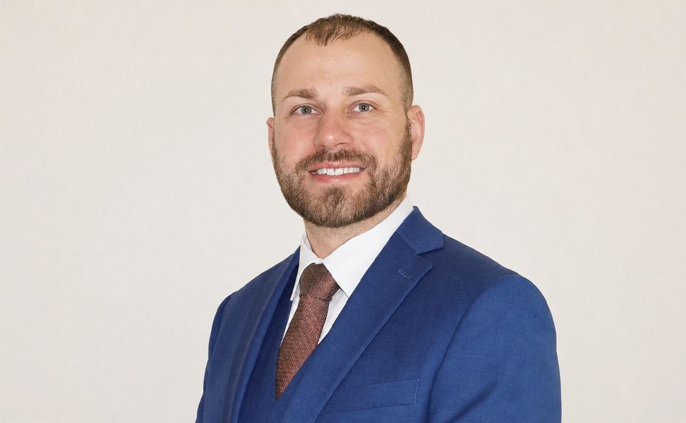 Jack Small wearing a blue suit and brown tie smiles against a plain background.