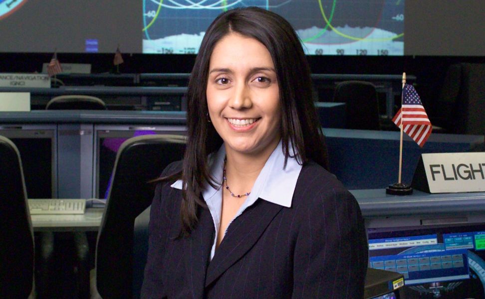 A woman in a suit smiling inside a control room, with monitors displaying charts and an American flag on desks in the background.