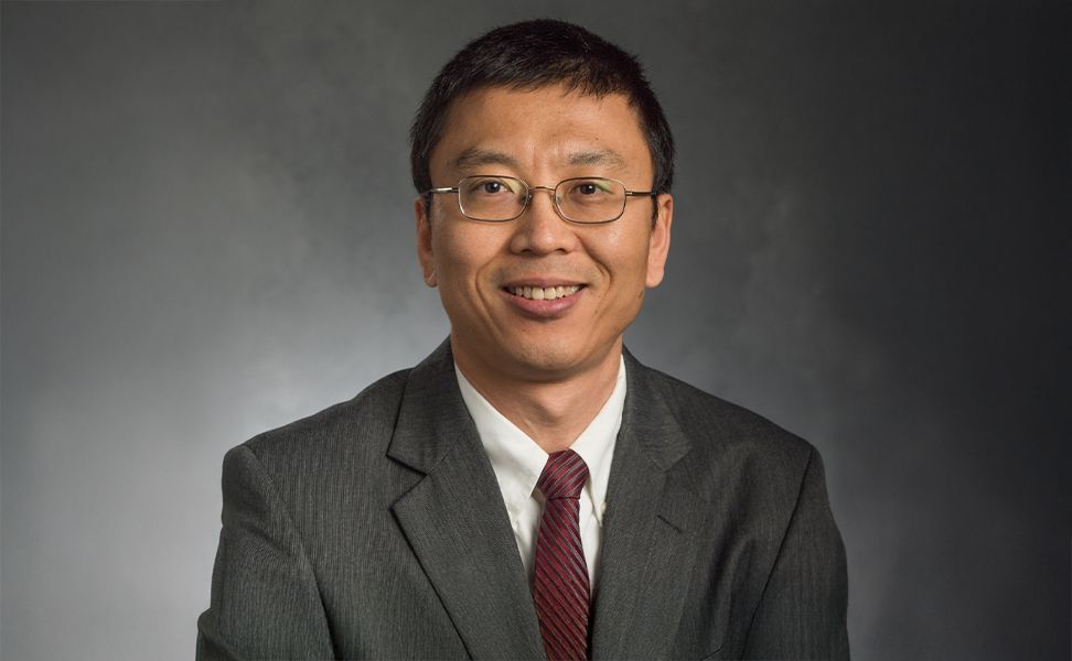 Dr. Ming Han in glasses wearing a gray suit and red striped tie, smiling at the camera against a dark gray background.