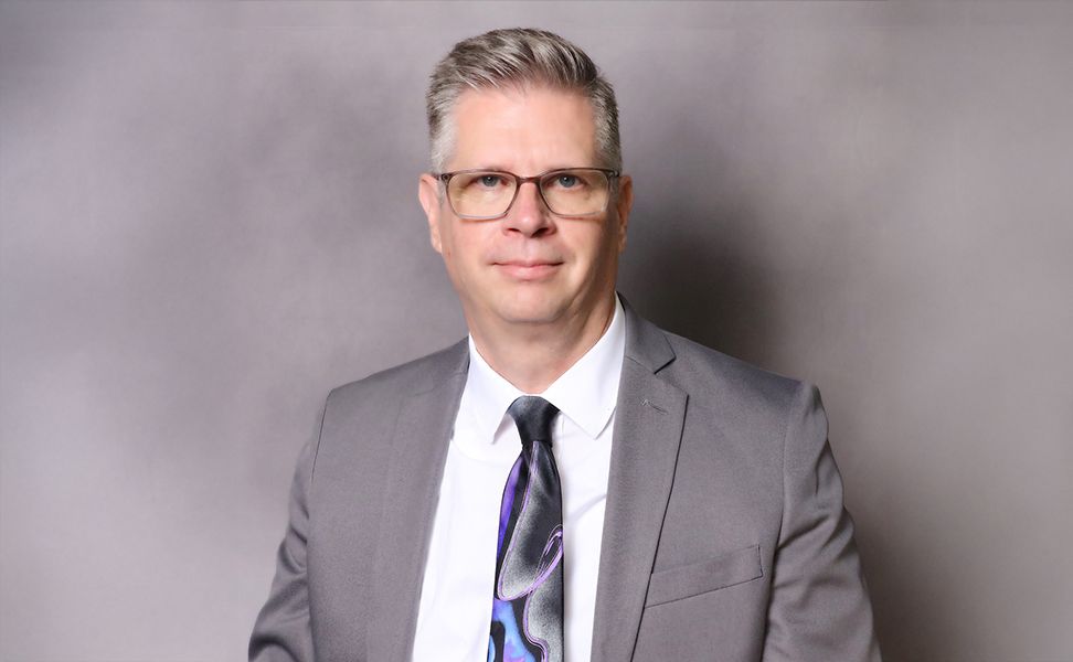 Barry Schieferstein, wearing a white shirt and a multicolored tie, against a neutral gray background.