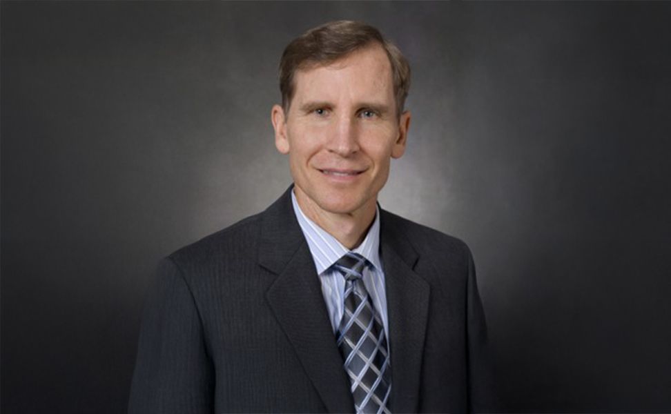 Dr. Gary Georgeson, in a suit and tie, smiling, against a dark, plain background.