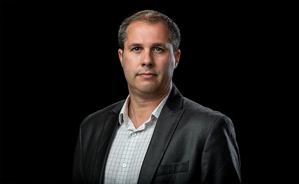 David Stobbe in a dark blazer and checkered shirt, with short hair, poses against a black background.