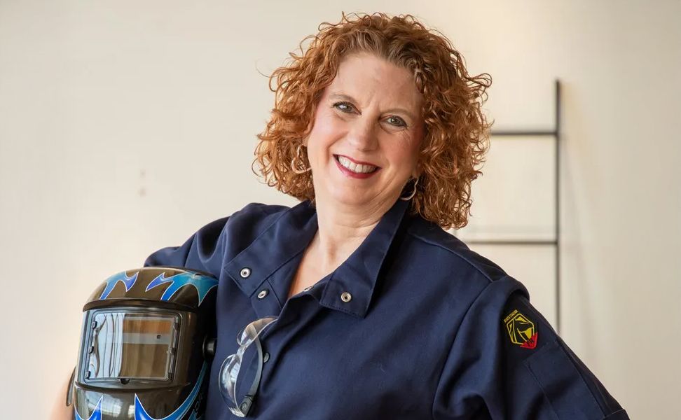 Smiling woman with curly hair wearing a navy blue jacket, holding a welding helmet with blue flames.