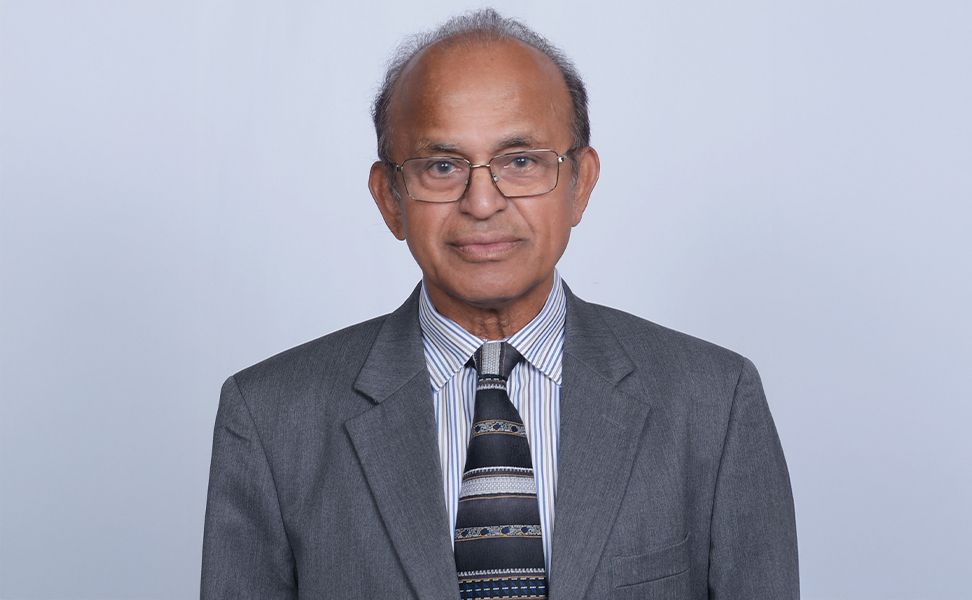 Ram Samy wearing a gray suit, striped shirt, and patterned tie, posing against a plain light background.