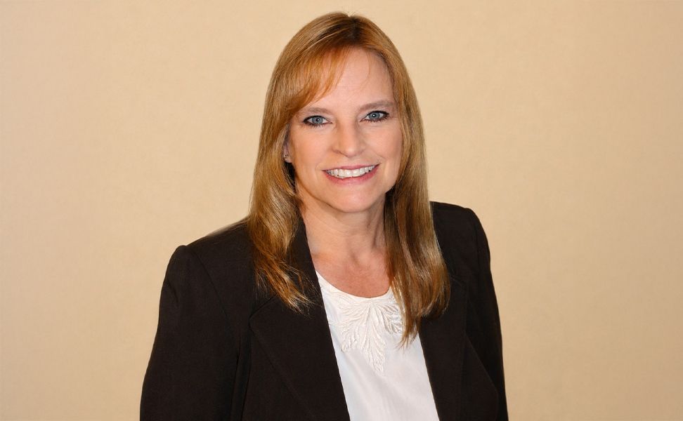 Jean Bridge, wearing a dark blazer and white blouse smiles against a light background.