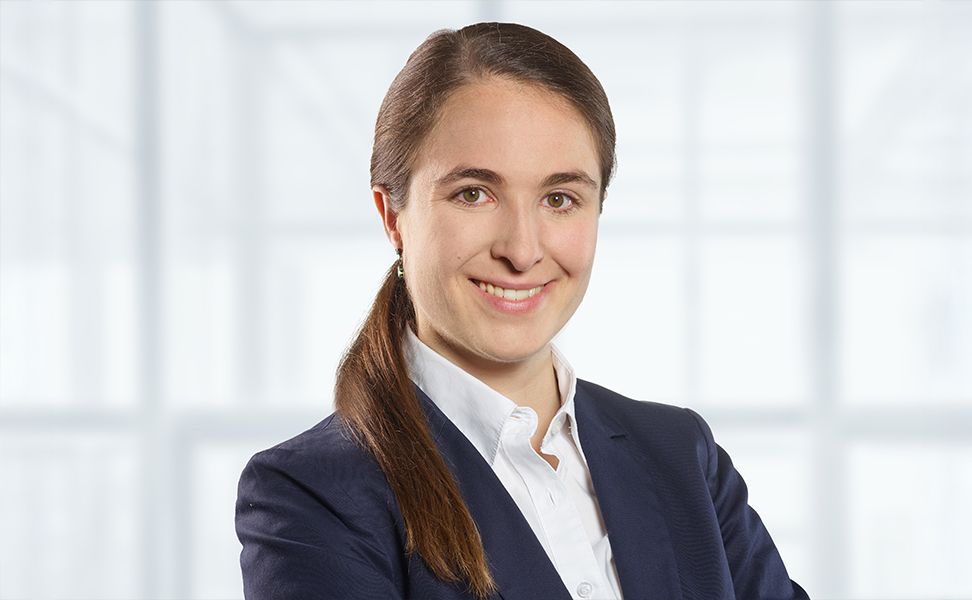 Sarah Fischer smiles at the camera with arms crossed, standing in a bright office with large windows in the background.