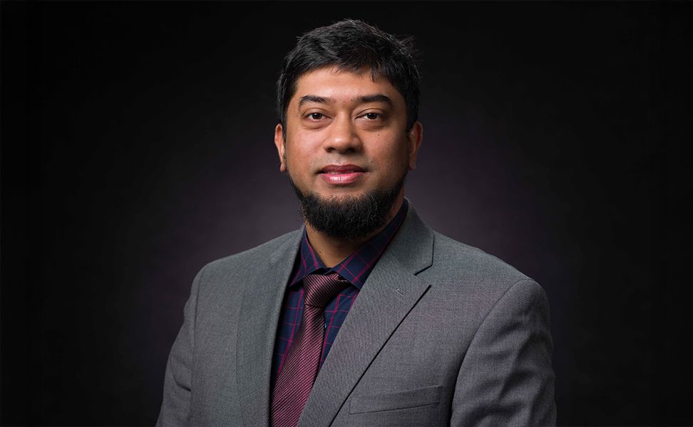 Dr. Showmic Islam in a grey suit and patterned shirt, wearing a tie, against a dark background.