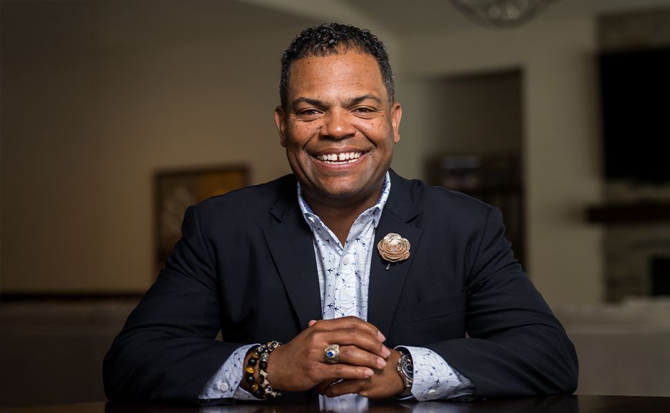 Jason O. Harris in a dark suit with a floral lapel pin, sits at a table with hands clasped, in a warmly lit room.