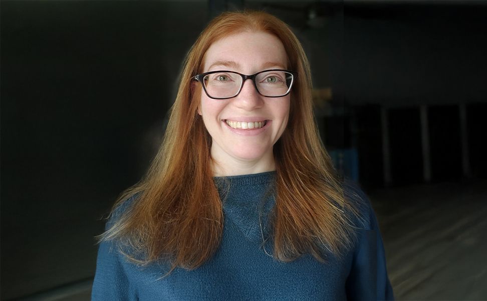Laura Homa, wearing a blue top, stands against a dark background.