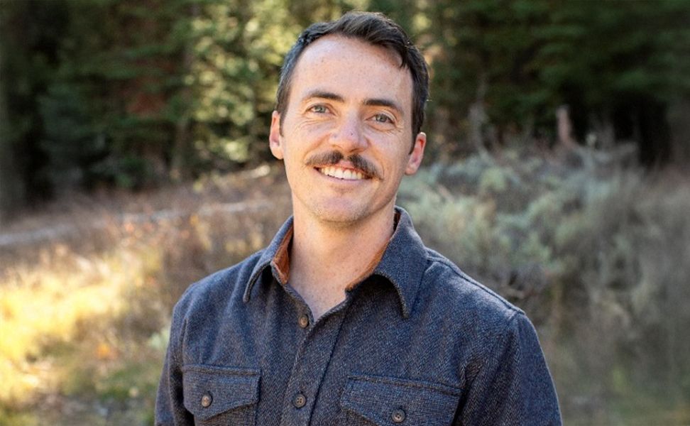 Taylor Sparks, smiling, wearing a blue shirt, stands outdoors with trees and grass in the background.