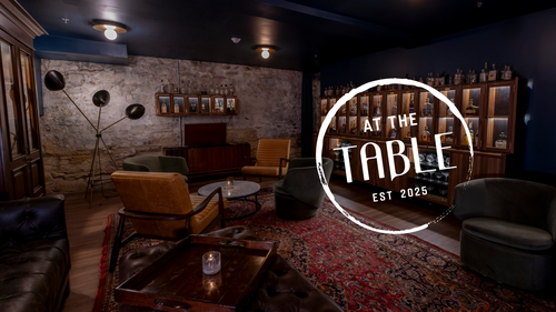 'At The Table' logo over photo of four chairs around a table in a dark, moody bar