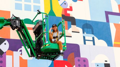 Alyssa in a lift in front of a large, colorful mural with abstract shapes and figures