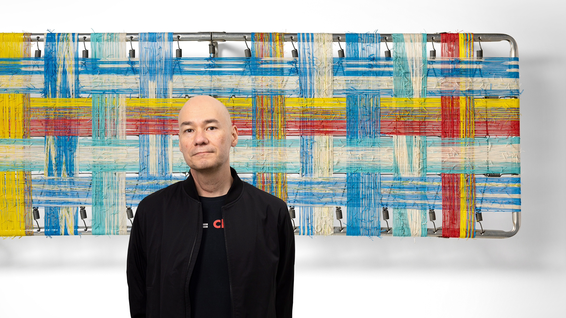 Brian stands in front of an abstract artwork with a colorful woven pattern of blue, red, yellow, and white strips.