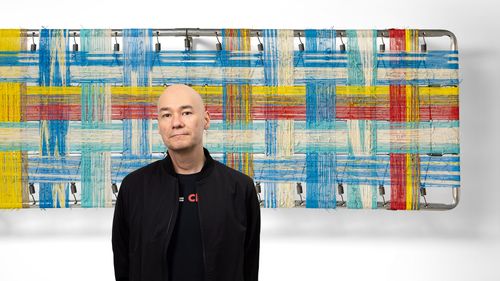 Brian stands in front of an abstract artwork with a colorful woven pattern of blue, red, yellow, and white strips.