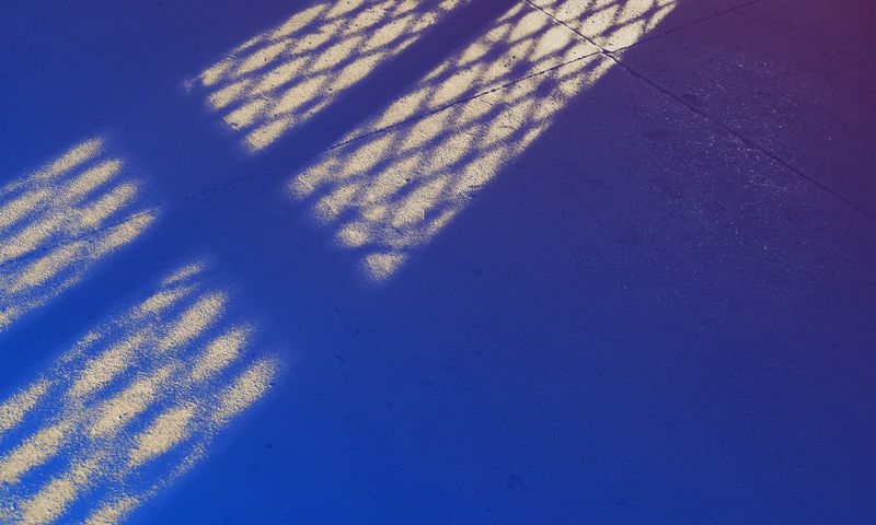 Patterned light shines through a lattice onto a smooth blue and purple floor, creating elongated, bright shadows across the surface.