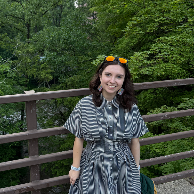 Tatiana in a striped dress wearing sunglasses, standing on a bridge in the middle of a forest