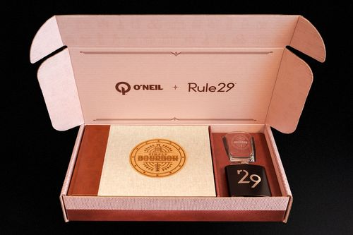 Opened promo box revealing O'Neil and Rule29 logos on the inside lid and the Simply bourbon book, bourbon glass, and R29 flask inside