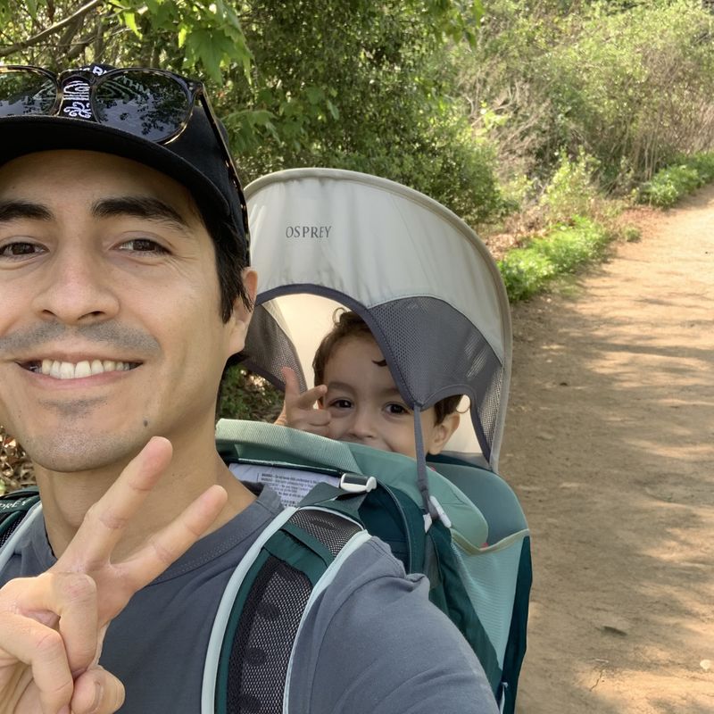 Edgar giving the peace sign during a hike, while wearing his child in a backpack carrier