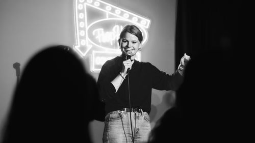 Nancy-O’Conner performs stand-up comedy, holding a microphone. A neon sign glows in the background. Black and white image.