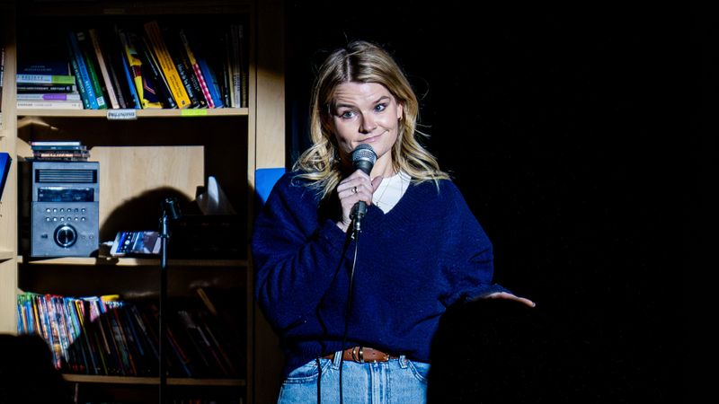 Nancy-O’Conner performs stand-up comedy, holding a microphone on a lit stage, with bookshelves in the background.