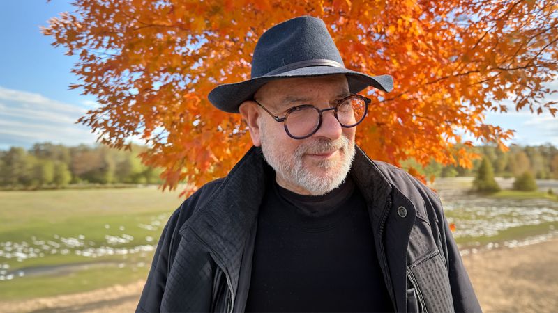 Steven Hellerstands in front of a vibrant orange autumn tree, with a grassy field in the background.