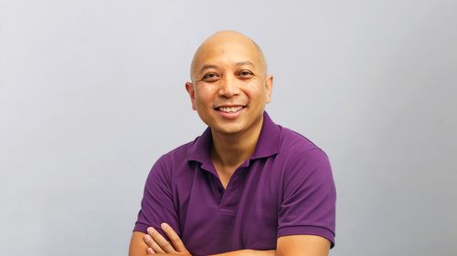 George Aye smiling, wearing a purple polo shirt, with arms crossed against a plain light gray background.