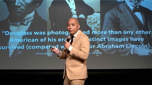 George Aye speaks with a microphone in front of a large screen displaying historical images and text.