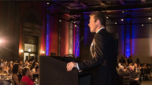 Jon Houghton speaks at a podium in an ornately decorated hall, addressing a seated audience. Purple and blue lights illuminate the pillars.