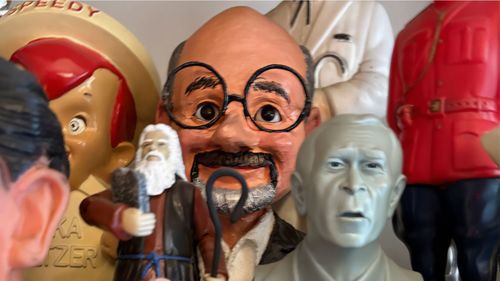 A collection of painted figurines and statues, including a bald man with large glasses, a bearded figure, and a silver bust.