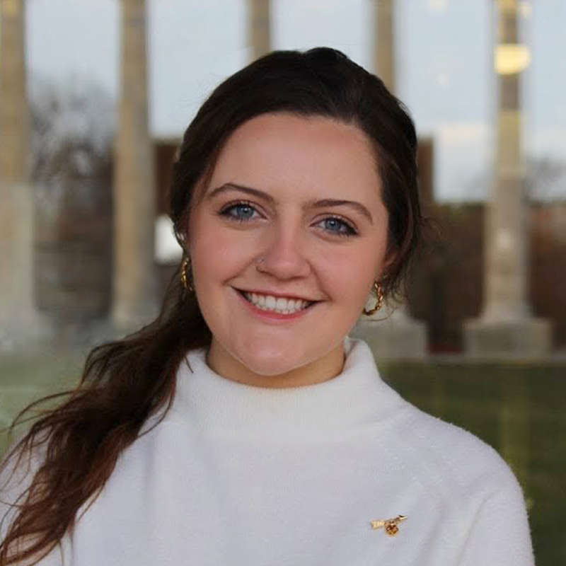 Lainey smiling outdoors in front of blurred columns and greenery