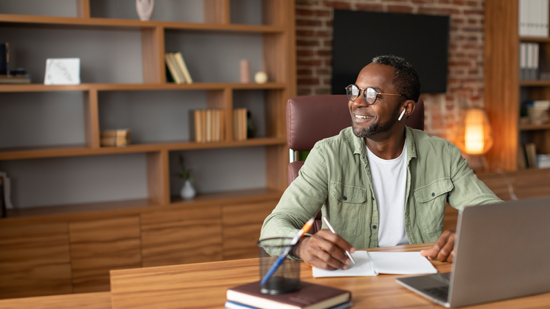 Man with glasses and earbuds sitting at a desk with a laptop and notebook.