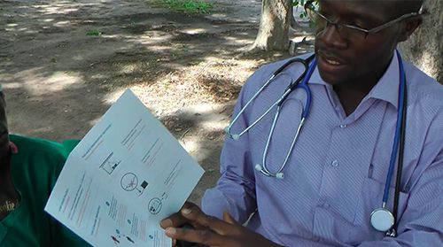 A person with a stethoscope explains a document with illustrations and text to someone outdoors under a tree.