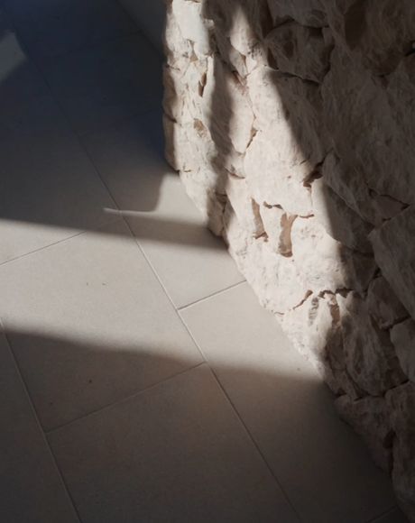 Sunlight casts shadows on a tiled floor and a textured stone wall, creating a play of light and shadow in a minimalist setting.