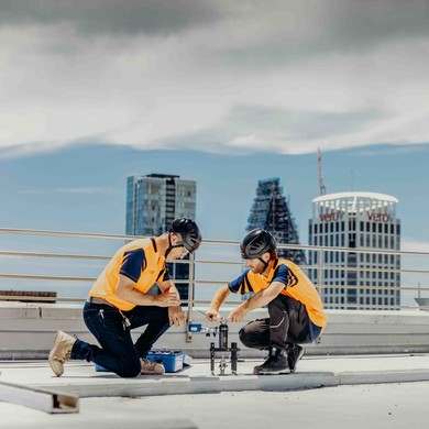 Two workers in install a fall arresst anchore on a rooftop, and test it for compliance