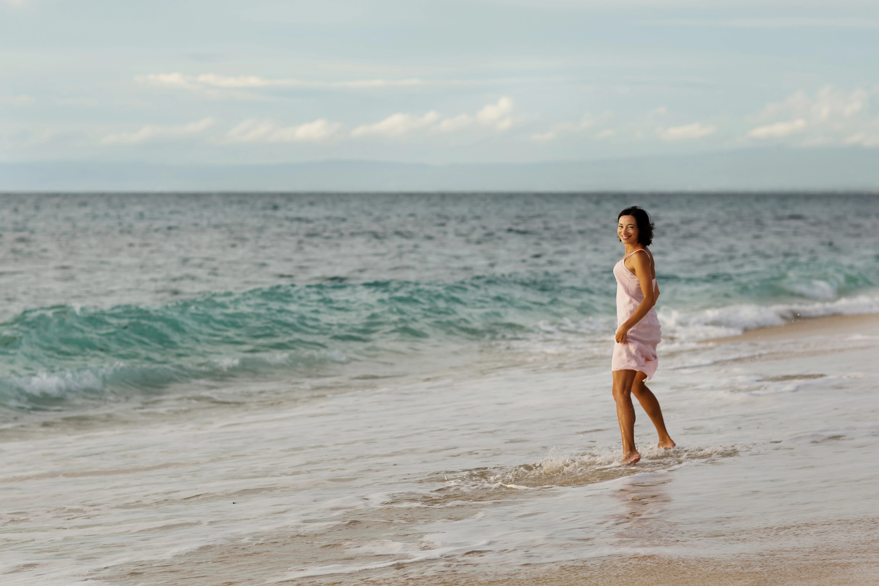 A person in a light dress walks along the shoreline of a tranquil beach, with gentle waves lapping at their feet and a cloudy sky in the background.