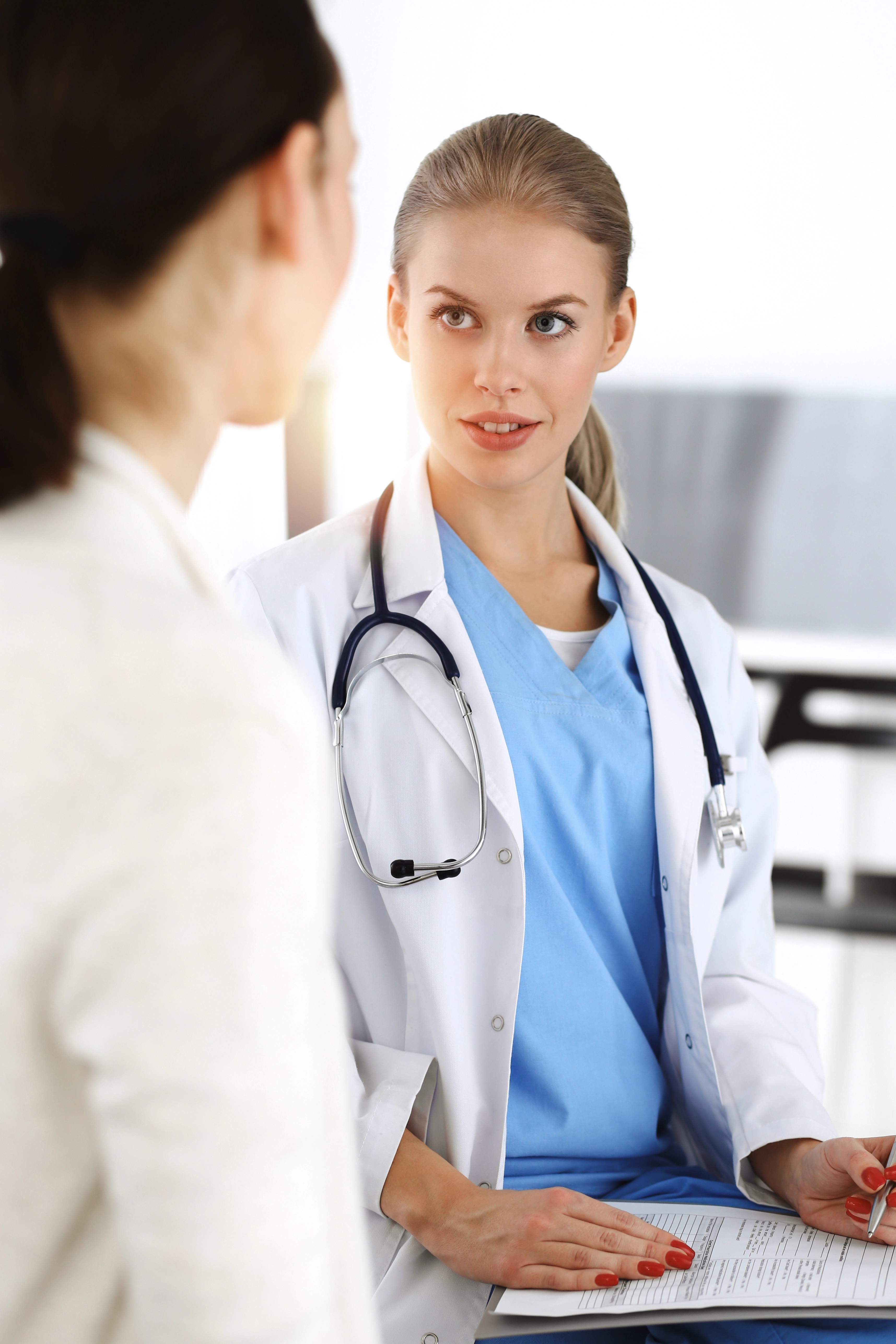 A female doctor in a white coat with a stethoscope talks to a patient while holding a clipboard.