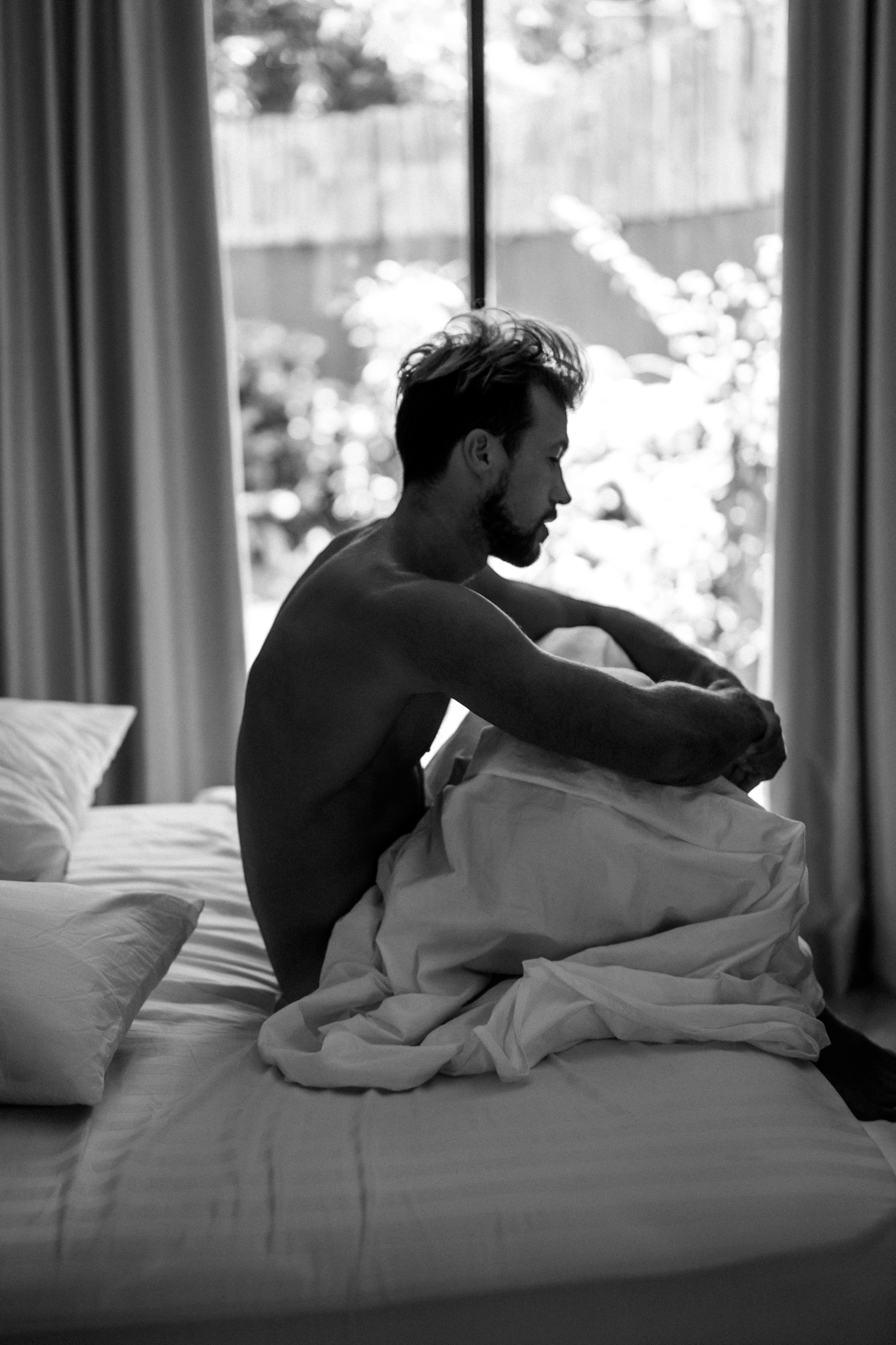 A black and white image of a man sitting on a bed with sheets, looking contemplative, near a window with soft light coming through.