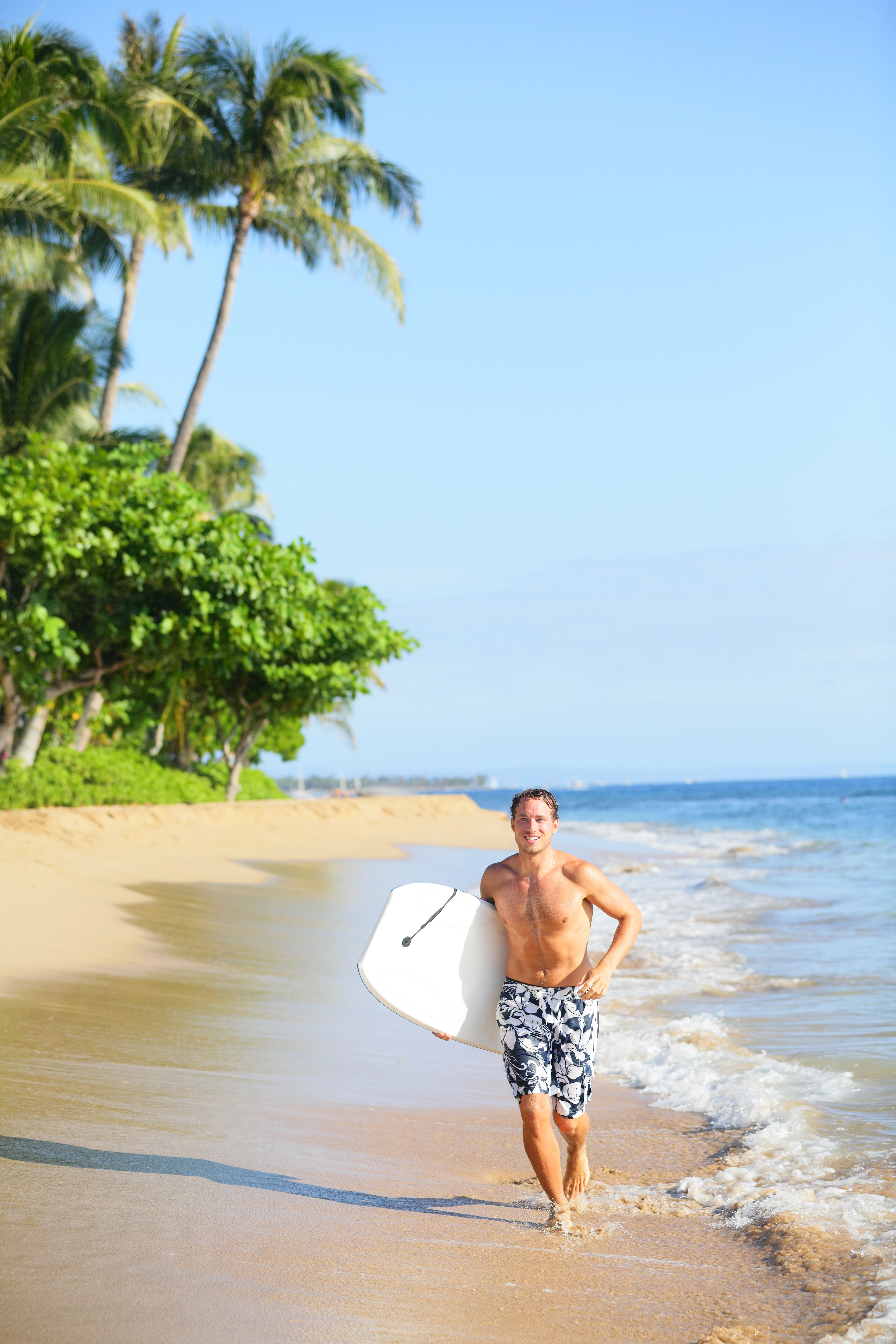 A man in swim trunks runs along a sandy beach, smiling and holding a bodyboard, with palm trees and ocean waves in the background.