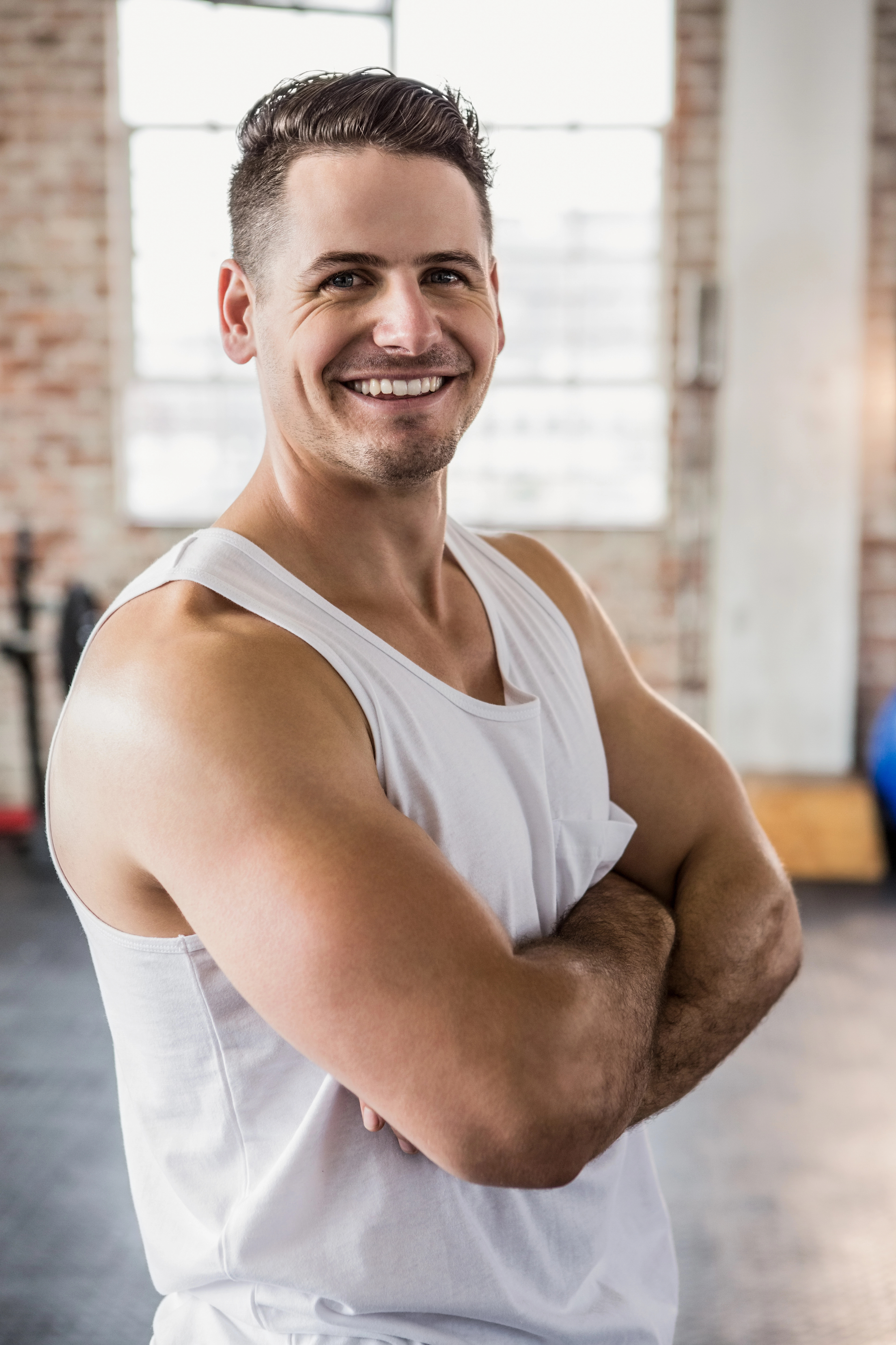 A man in a white tank top stands confidently with arms crossed, smiling in a well-lit gym with exposed brick walls and fitness equipment.