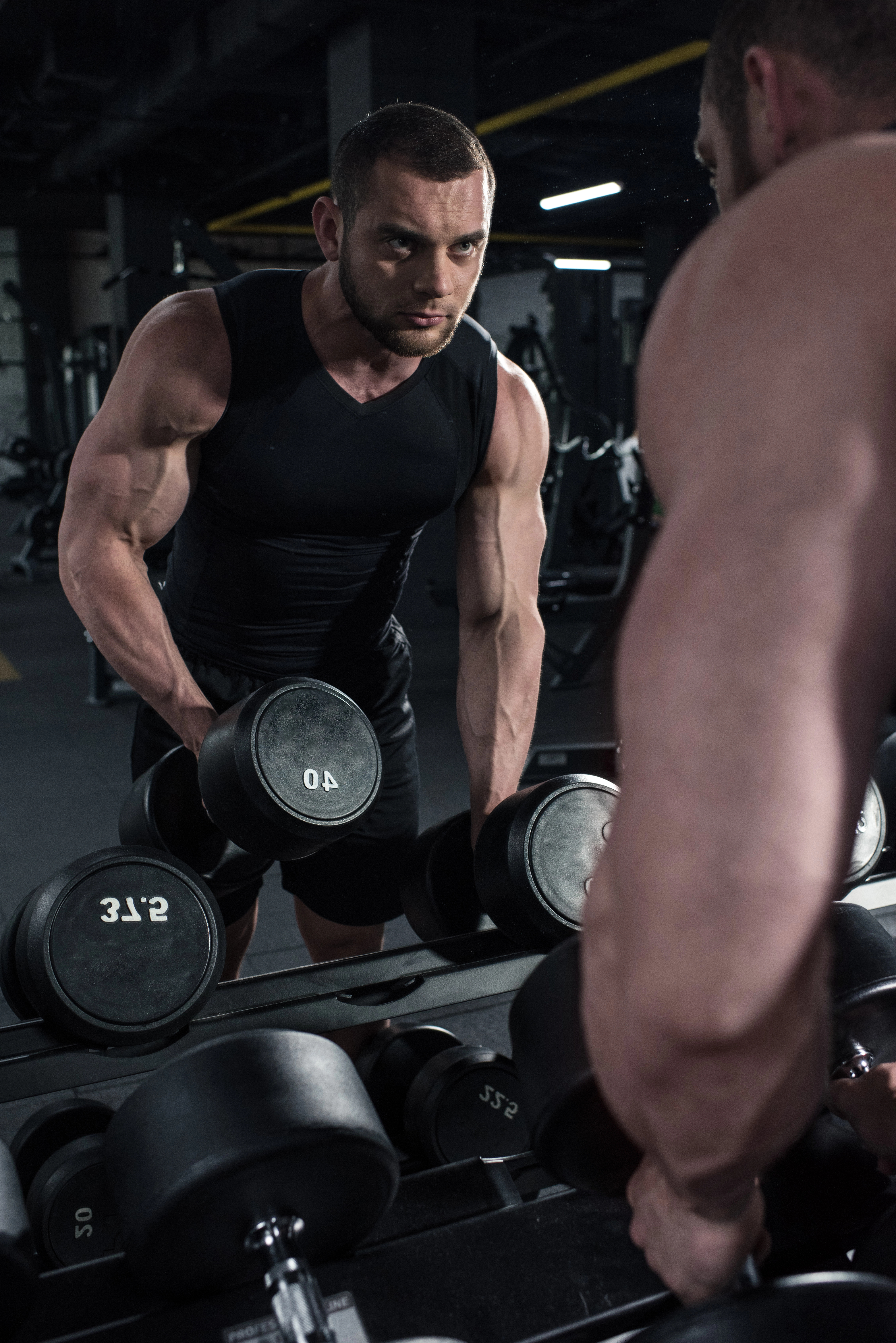 Man in a gym wearing a black tank top, holding a 40-pound dumbbell, looking intensely at his reflection in the mirror.