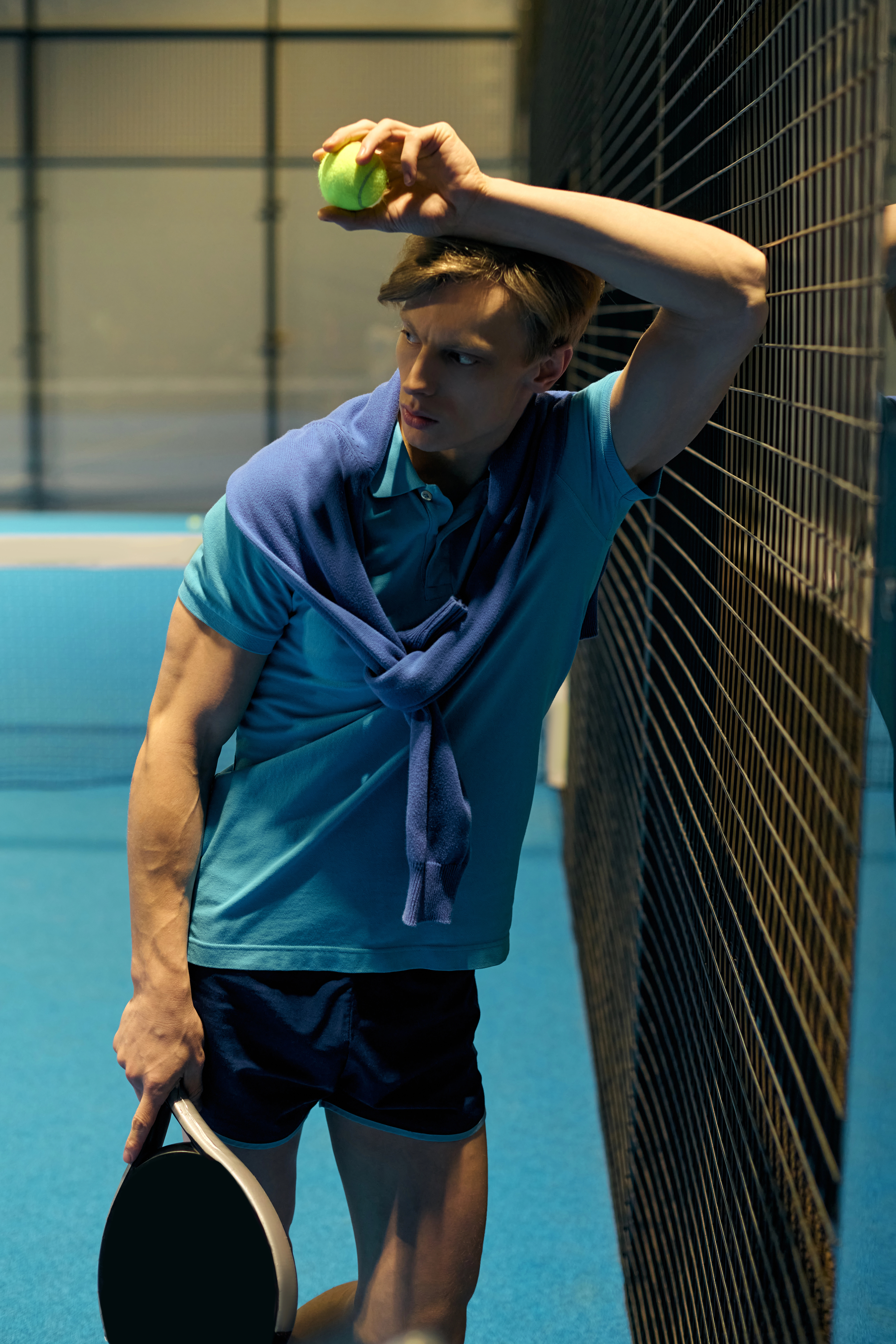 Man in a blue shirt and sweater rests against a fence, holding a tennis ball and paddle, with a focused expression in an indoor court.