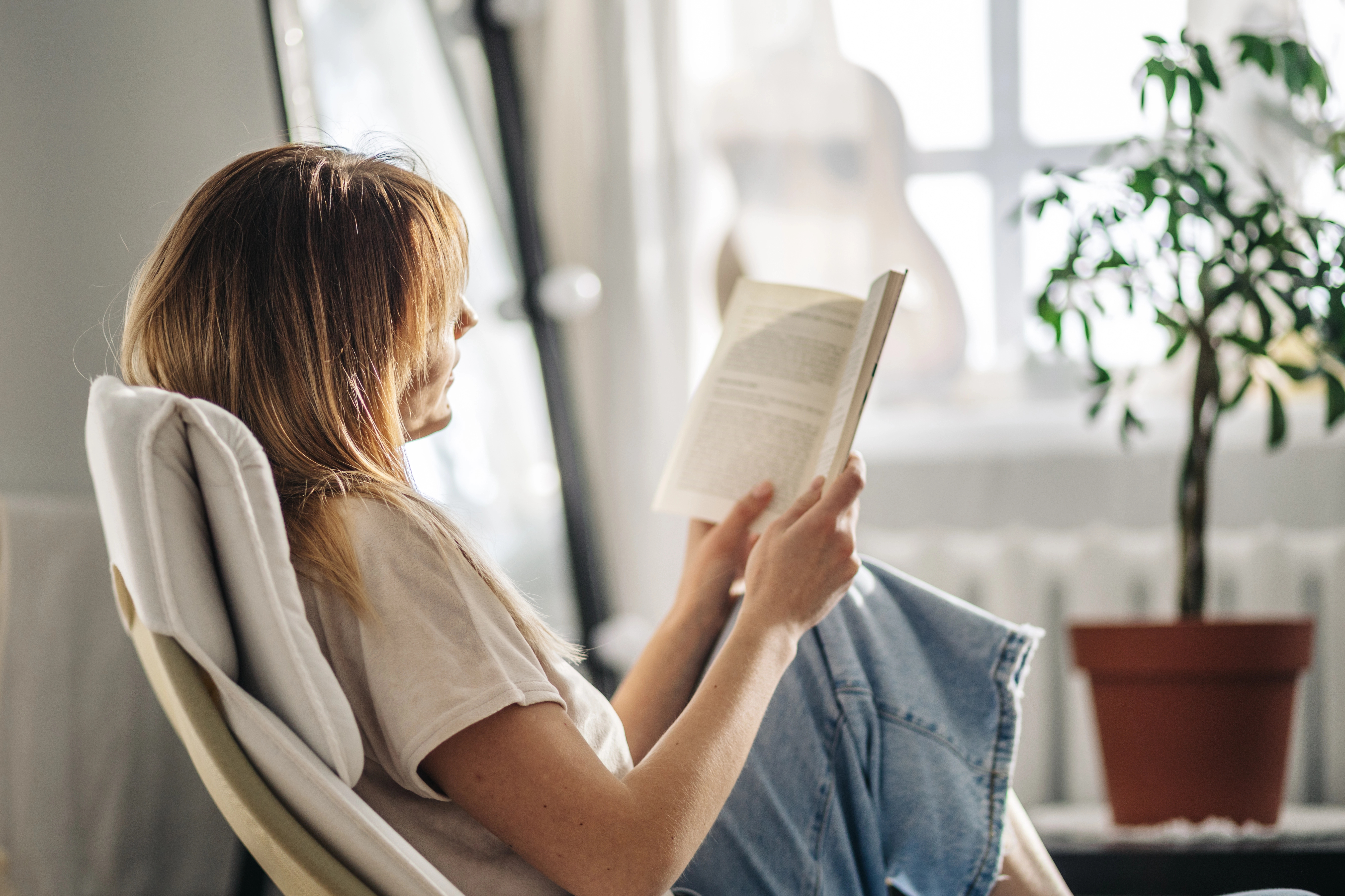 A woman with long hair, wearing a white shirt, sits in a chair reading a book near a window with a potted plant nearby.