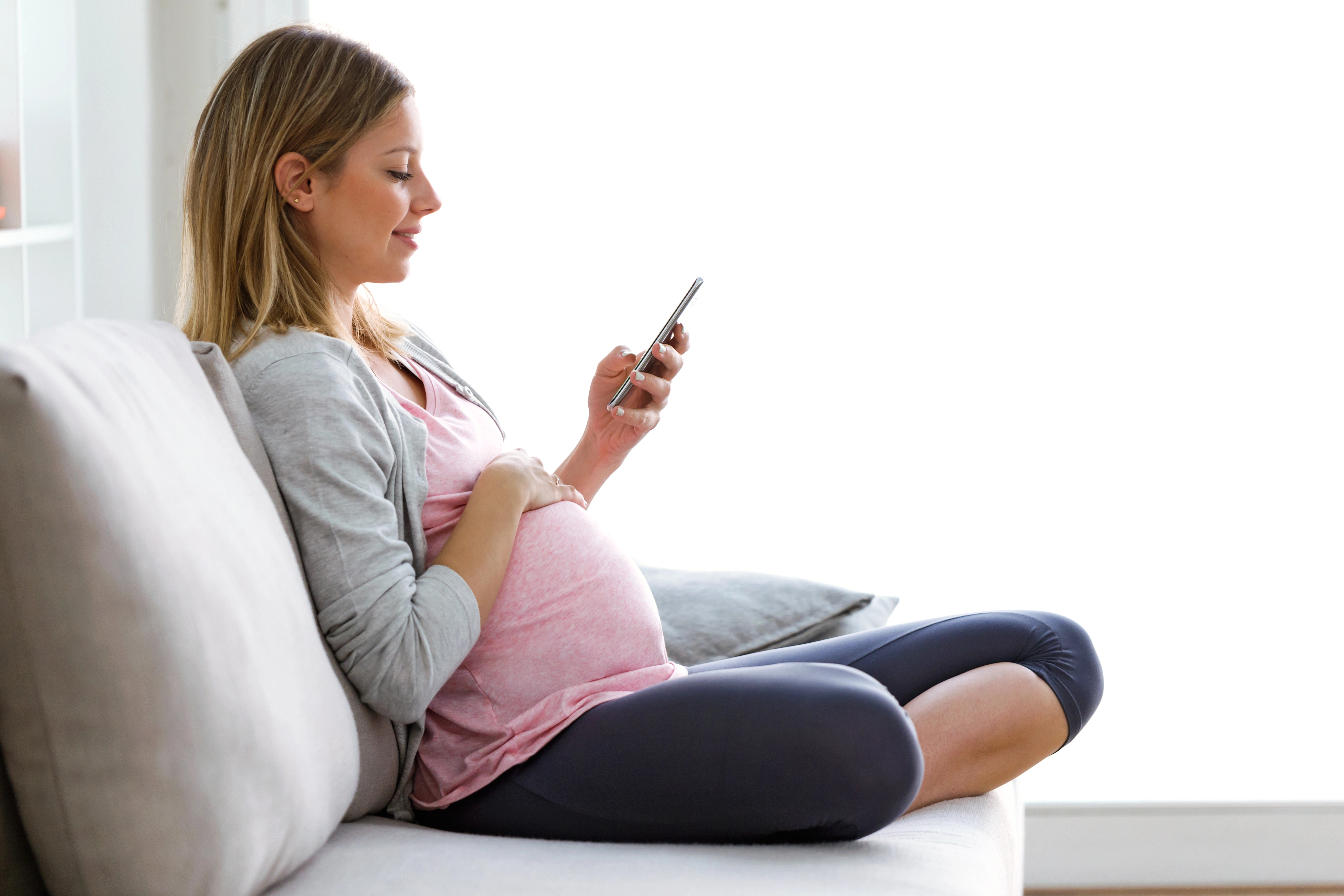 Pregnant woman sitting on a couch, smiling while looking at her smartphone and resting her hand on her belly.