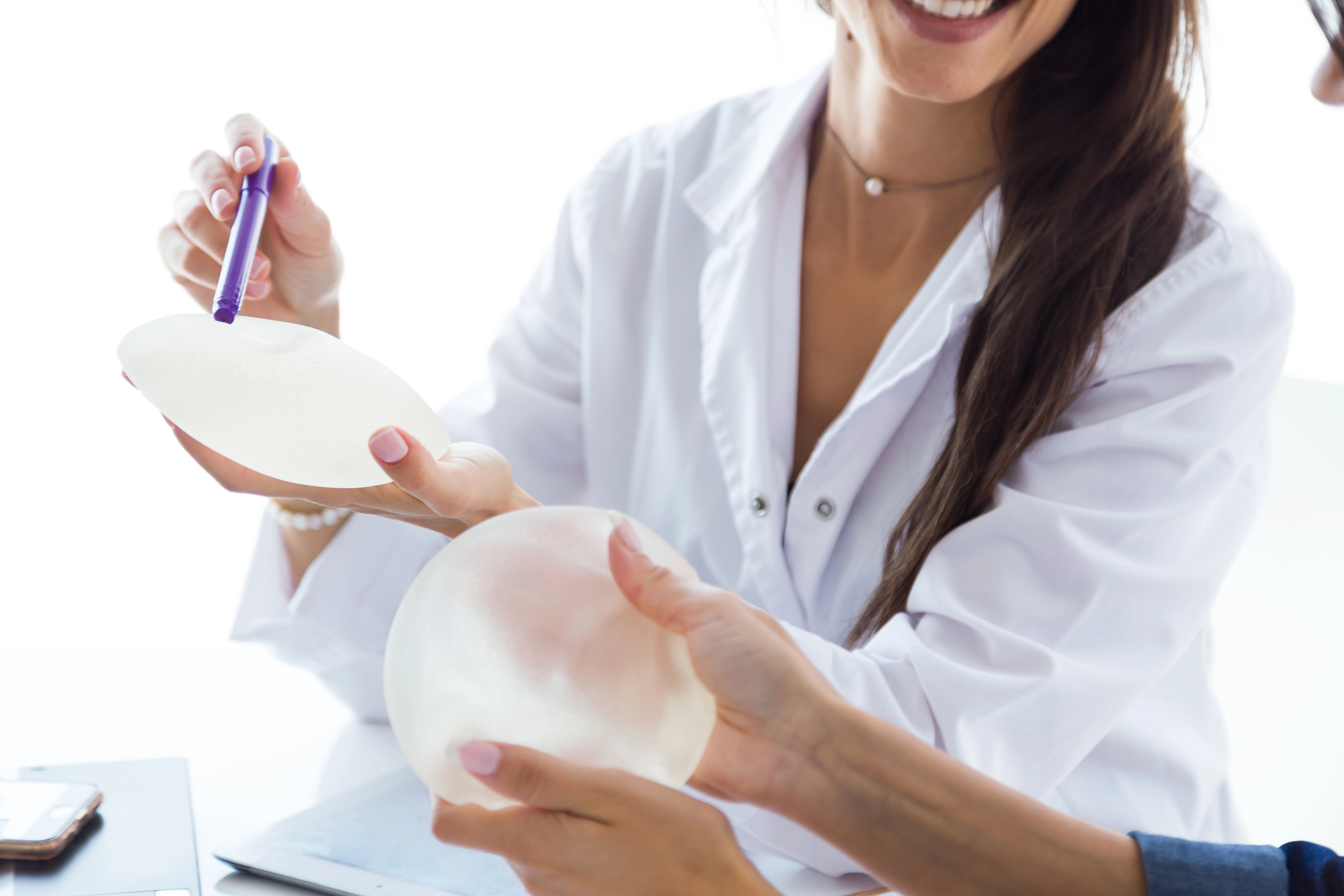 Two women in lab coats examining breast implants, one holding an implant while the other points with a pen; smiles and discussion visible.