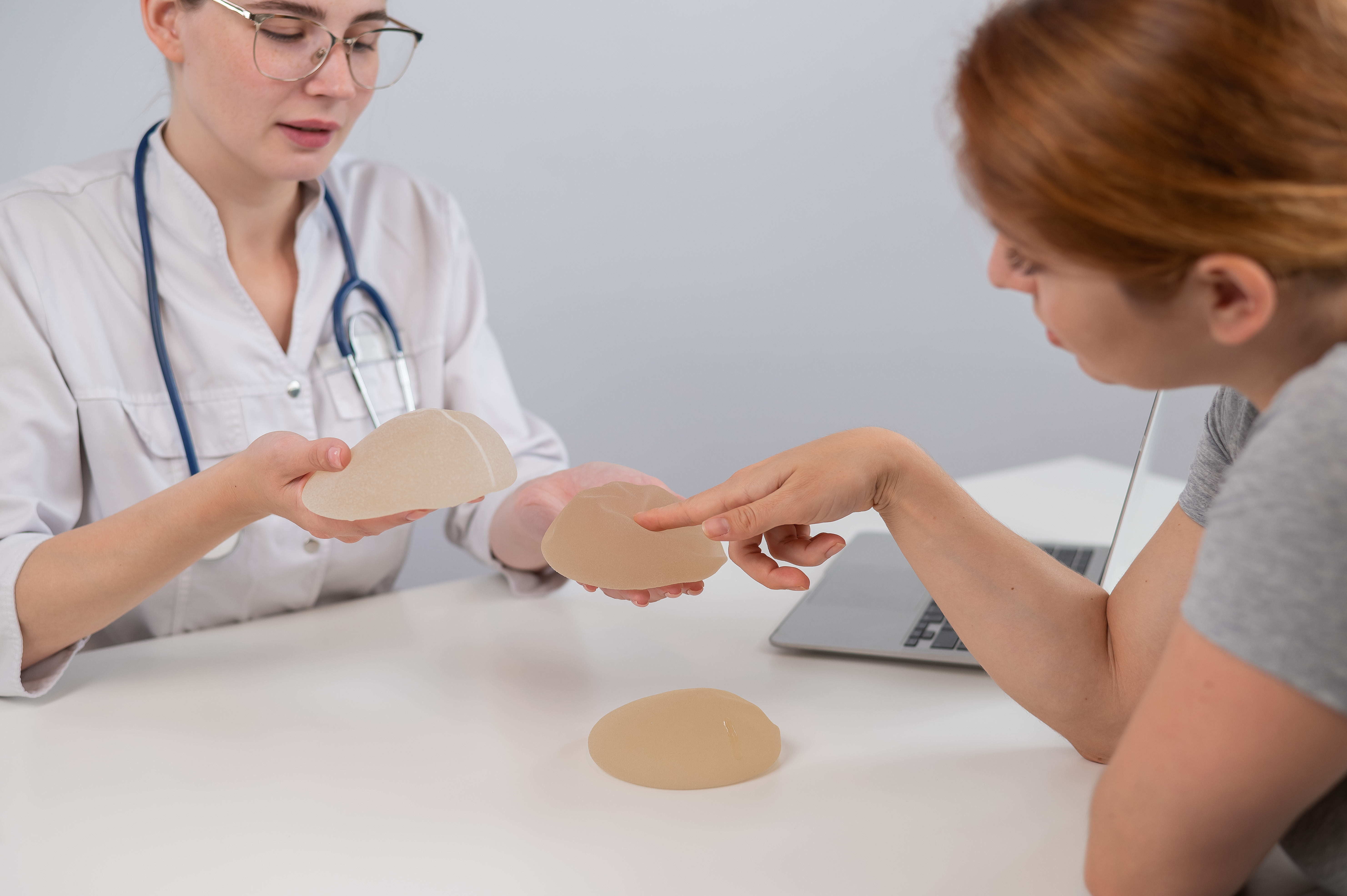 Doctor discussing breast implants with a patient at a desk, showing different implant options. The patient is pointing at an implant.