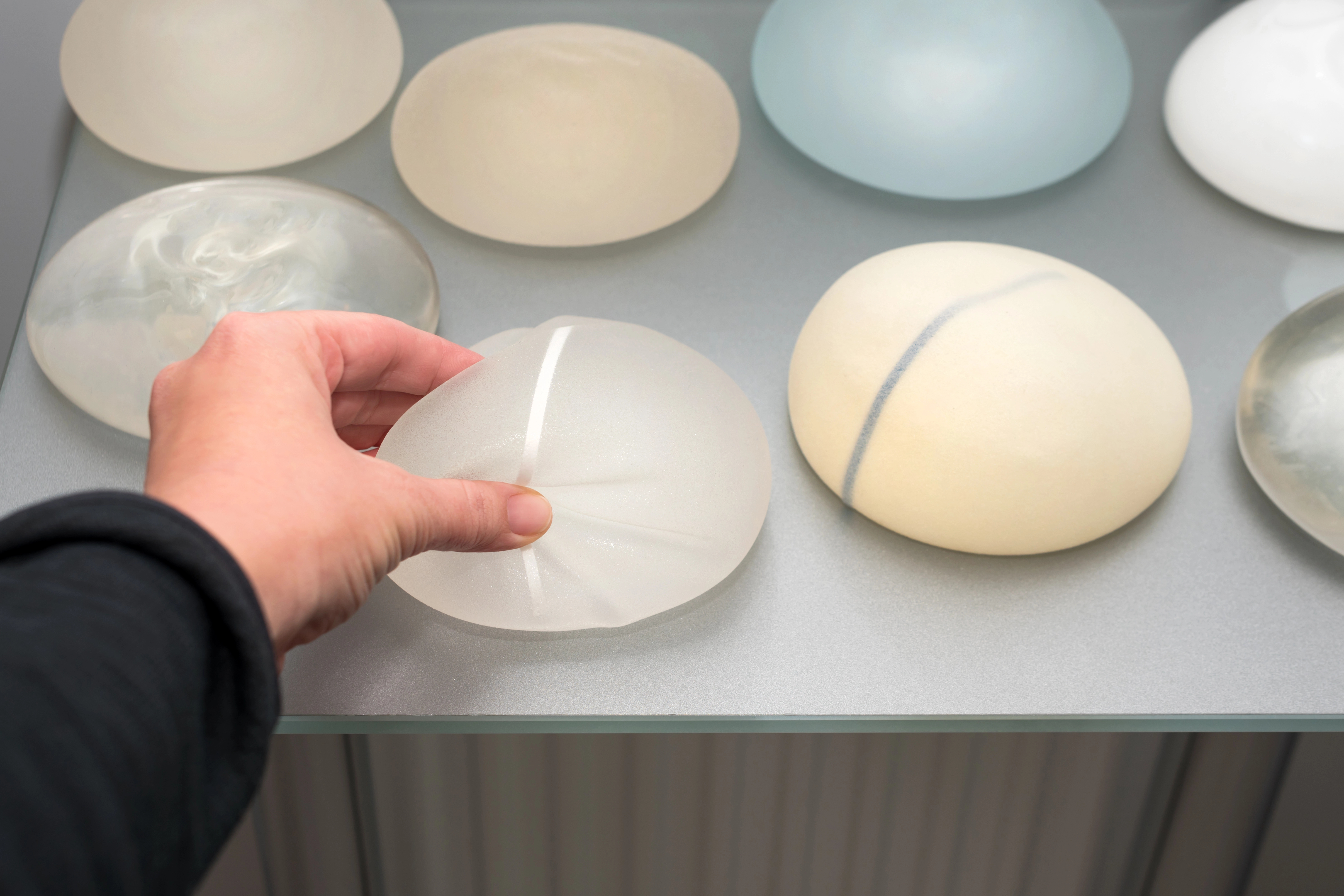 A hand touching a silicone breast implant on a table with several other implants of varying shapes and colors displayed.