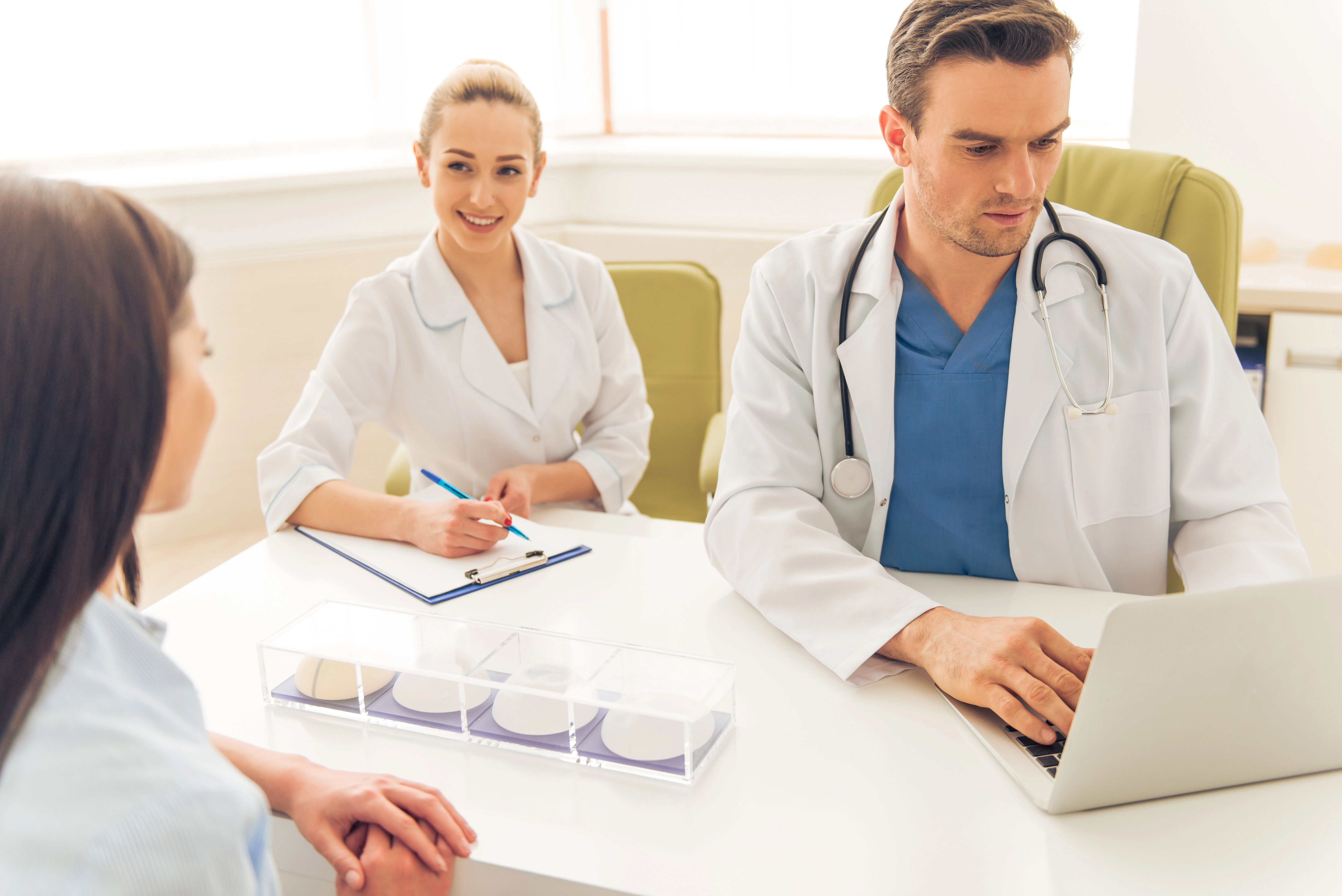 Doctors in consultation with a patient. The male doctor is using a laptop, while the female doctor takes notes. Breast implants are on the table.