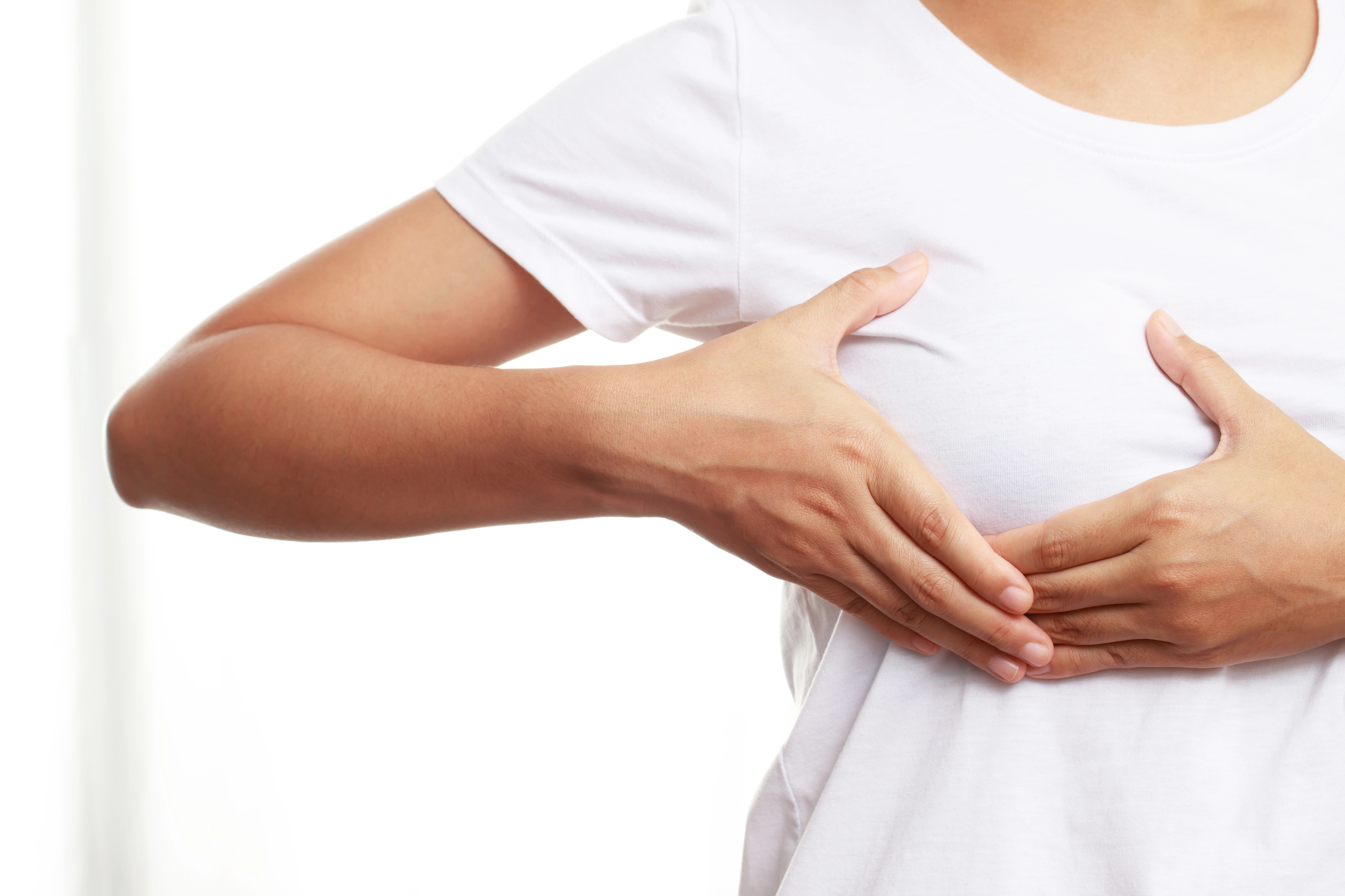 Person in a white shirt performing a breast self-exam by touching their chest with both hands against a light background.