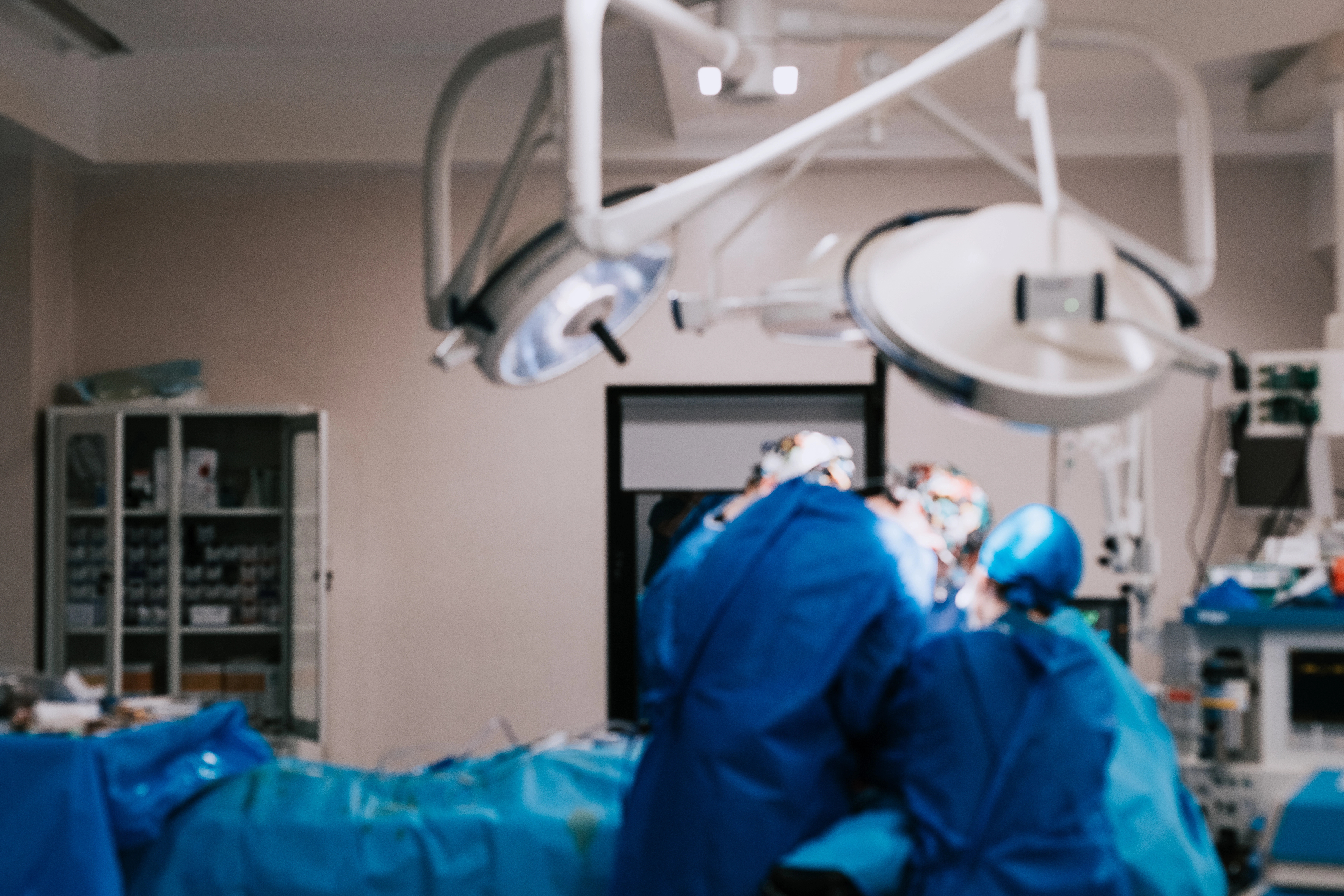 Surgeons in blue scrubs and caps perform an operation under bright surgical lights in an operating room.